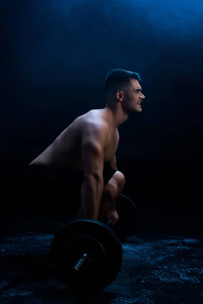 Side view of sexy muscular bodybuilder with bare torso excising with barbell on black background with smoke — Stock Photo