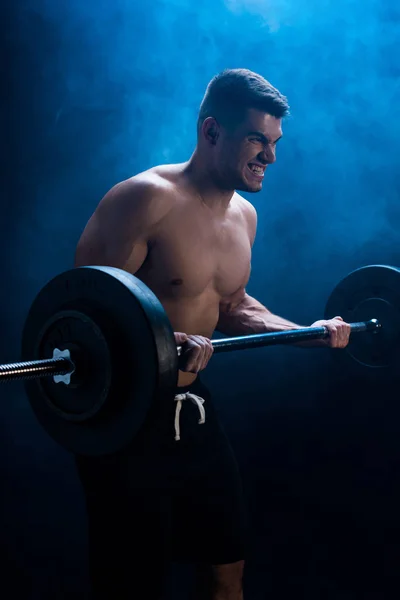 Tense muscular bodybuilder with bare torso excising with barbell on black background with smoke — Stock Photo