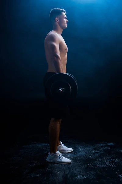 Vue latérale du bodybuilder musclé sexy avec torse nu excisant avec haltère sur fond noir avec fumée — Photo de stock