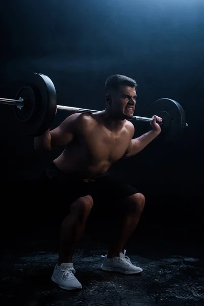 Tense sexy muscular bodybuilder with bare torso squatting with barbell on black background — Stock Photo