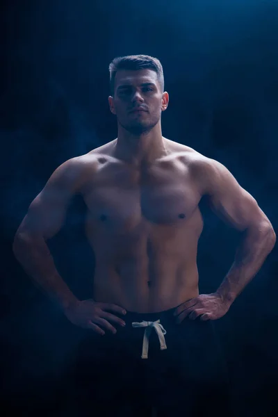 Sexy muscular bodybuilder with bare torso posing with hands on hips on black background with smoke — Stock Photo