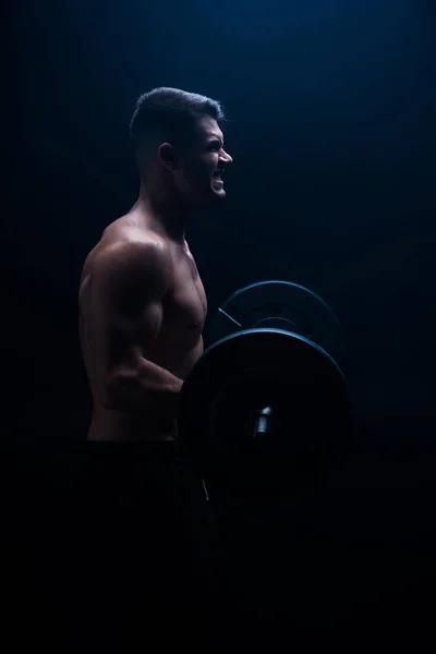 Seitenansicht von sexy muskulösen Bodybuilder mit nacktem Oberkörper beim Schneiden mit Langhantel auf schwarzem Hintergrund — Stockfoto