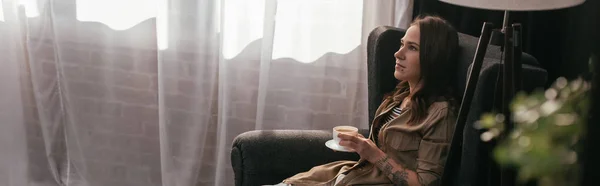 Tiro panorâmico de mulher bonita segurando xícara de café enquanto sentado em poltrona em casa — Fotografia de Stock