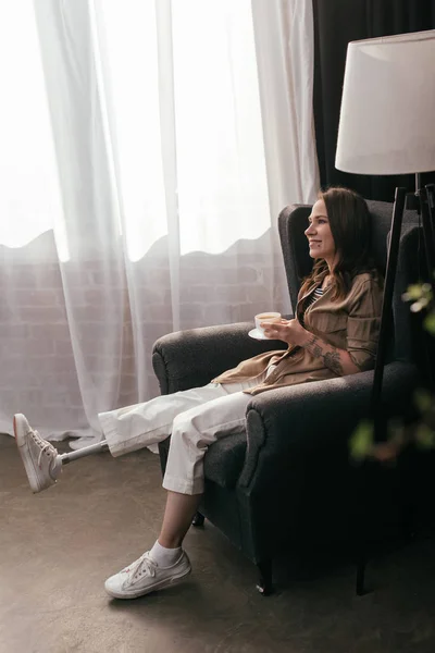 Вид сбоку красивой улыбающейся женщины с протезом ноги, которая держит кофе, сидя в кресле — стоковое фото
