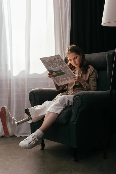 Menina com prótese perna ler jornal em poltrona em casa — Fotografia de Stock