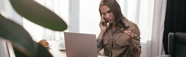 Enfoque selectivo de la mujer joven hablando en el teléfono inteligente y mirando el ordenador portátil en la cocina, plano panorámico - foto de stock