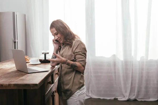 Vue latérale d'une femme souriante parlant sur un smartphone à côté d'un ordinateur portable sur une table de cuisine — Photo de stock