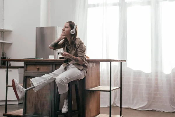 Hermosa mujer joven con la pierna protésica usando auriculares y teléfono inteligente en la cocina - foto de stock