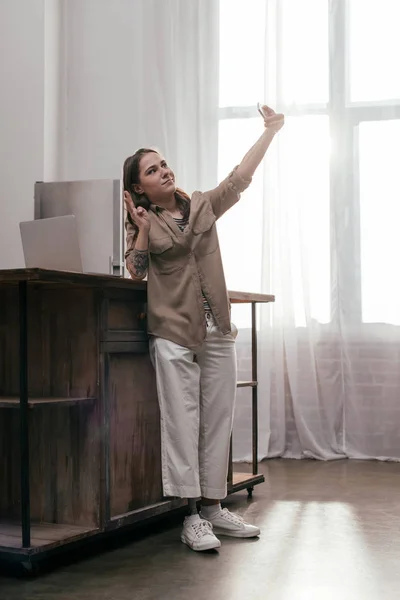 Молода жінка з протезною ногою бере селфі зі знаком миру біля ноутбука на кухонному столі — стокове фото