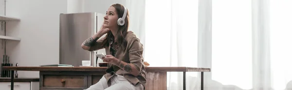 Tiro panorâmico de jovem mulher em fones de ouvido segurando smartphone na cozinha — Fotografia de Stock