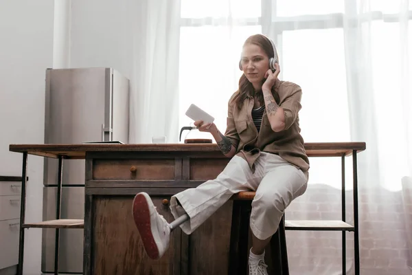 Молода жінка з протезною ногою використовує навушники і тримає смартфон, щоб бути кухонним столом — стокове фото