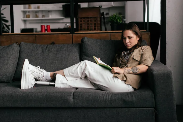 Mujer joven con prótesis de pierna libro de lectura en el sofá en la sala de estar - foto de stock