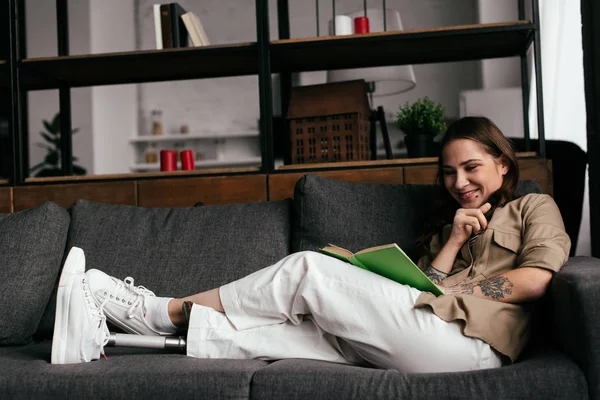 Mujer sonriente con prótesis de pierna libro de lectura en el sofá en la sala de estar - foto de stock