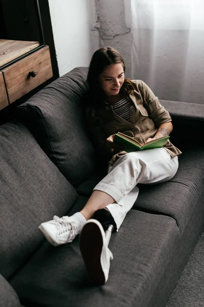 High angle view of smiling woman with prosthetic leg reading book on sofa at home — Stock Photo