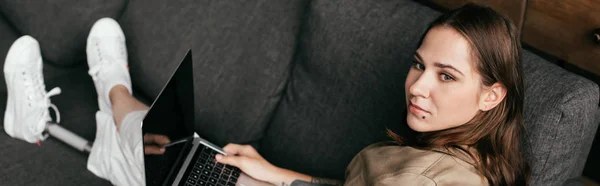 Selective focus of woman with prosthetic leg using laptop with blank screen on sofa, panoramic shot — Stock Photo