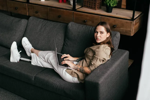 Jeune femme avec prothèse jambe tenant ordinateur portable et regardant loin sur le canapé — Photo de stock