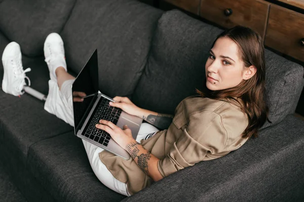 Hermosa chica con la pierna protésica utilizando el ordenador portátil con pantalla en blanco y mirando hacia otro lado en el sofá - foto de stock