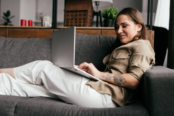 Вид збоку усміхненої жінки, що використовує ноутбук на дивані у вітальні — стокове фото