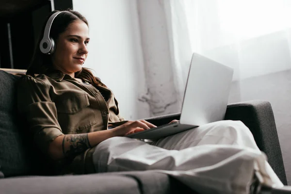 Selective focus of smiling girl with leg prosthesis using laptop and headphones on sofa — Stock Photo