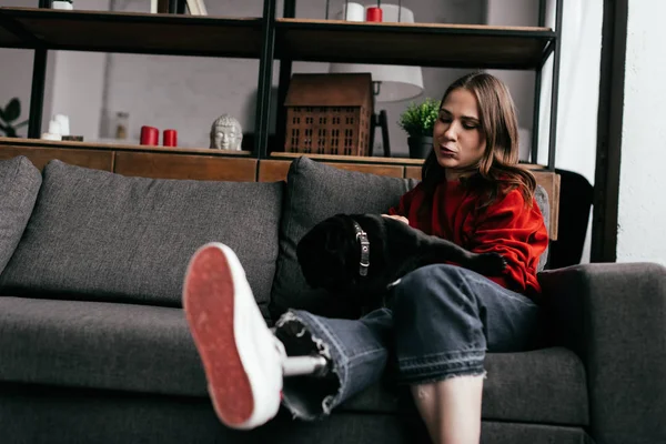 Selective focus of young woman with prosthetic leg petting pug dog on sofa in living room — Stock Photo