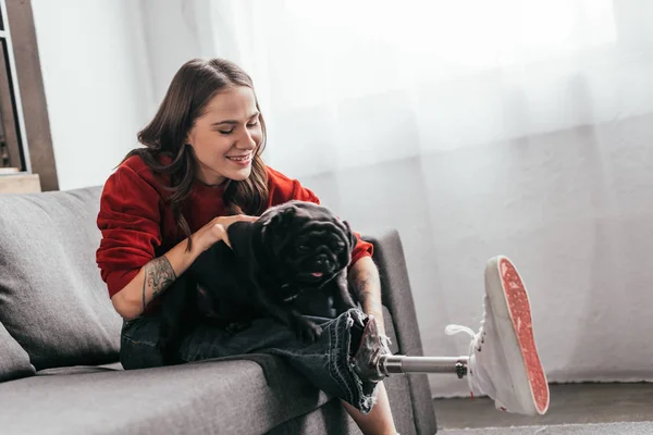 Smiling woman with prosthetic leg playing with pug on sofa — Stock Photo