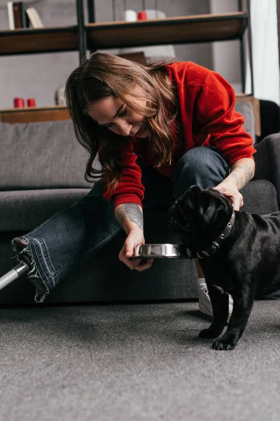 Young woman with prosthetic leg feeding pug by sofa in living room — Stock Photo