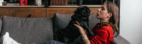 Seitenansicht der attraktiven Frau, die mit Mops auf Sofa spielt, Panoramaaufnahme — Stockfoto