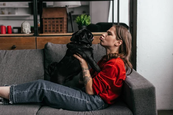 Side view of attractive woman grimacing to pug dog on sofa — Stock Photo