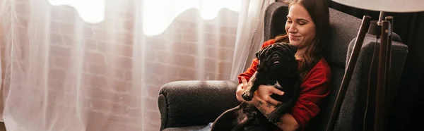 Усміхнена дівчина тримає мопса собаку в кріслі вдома, панорамний постріл — стокове фото