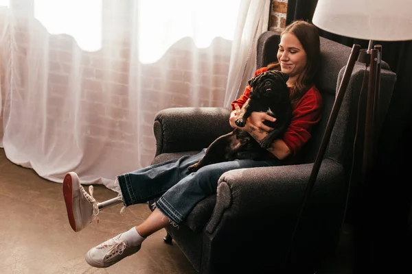 Усміхнена жінка з протезною ногою тримає мопса собаку в кріслі вдома, панорамний знімок — стокове фото