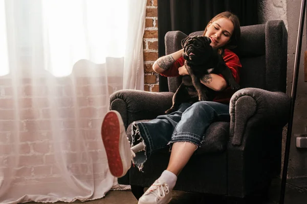 Lächelnde junge Frau mit Beinprothese spielt zu Hause mit Mops im Sessel — Stockfoto