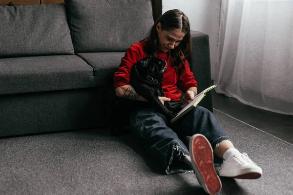 Woman with leg prosthesis reading book and holding pug dog on floor in living room — Stock Photo