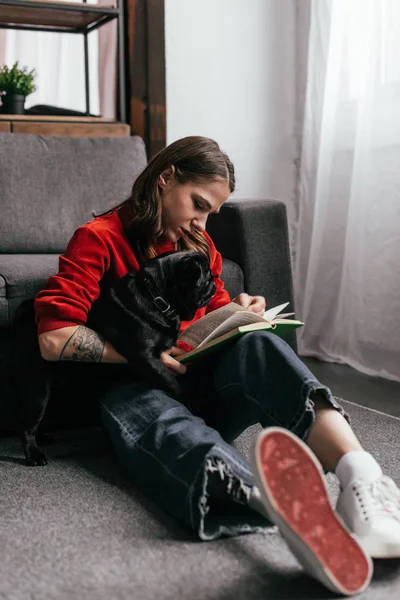 Enfoque selectivo de chica con libro de lectura de piernas protésicas y pug abrazo en el suelo en la sala de estar - foto de stock