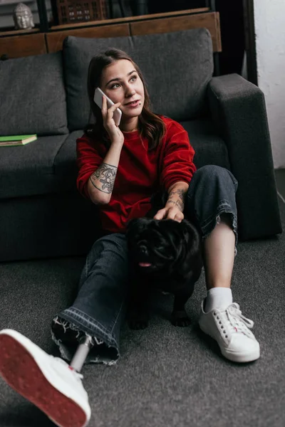 Frau mit Beinprothese spricht auf Smartphone, während sie Mops im Wohnzimmer auf dem Boden streichelt — Stockfoto