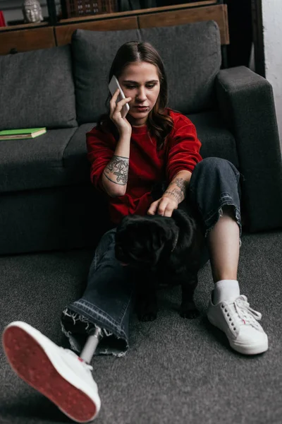 Mujer con la pierna protésica hablando en el teléfono inteligente mientras acaricia pug en el suelo en la sala de estar - foto de stock