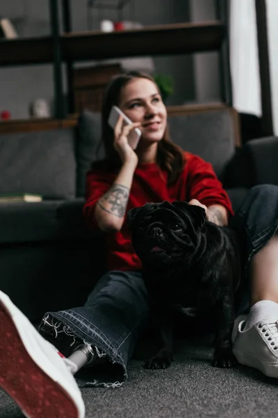 Foco seletivo de mulher sorridente com perna protética falando no smartphone ao lado de pug no chão na sala de estar — Fotografia de Stock