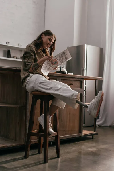 Mujer joven sonriente con prótesis de pierna leyendo periódico en la cocina - foto de stock