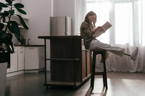 Vista lateral de mujer con pierna protésica leyendo periódico por mesa de cocina - foto de stock