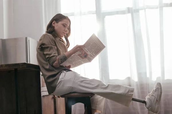 Vista lateral de mujer con pierna protésica leyendo periódico por mesa de cocina - foto de stock