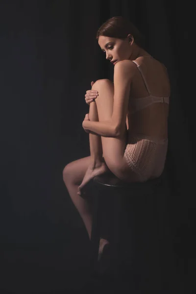 Schöne junge Frau in Dessous posiert, während sie auf schwarzem Hintergrund sitzt — Stockfoto