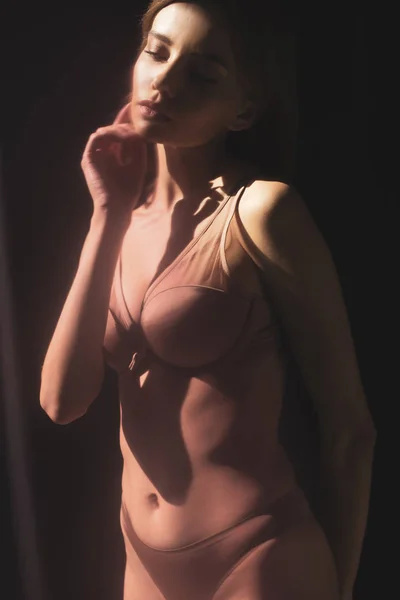 Сексуальна дівчина в нижній білизні торкається обличчя, позує на сонячному світлі на чорному фоні — стокове фото
