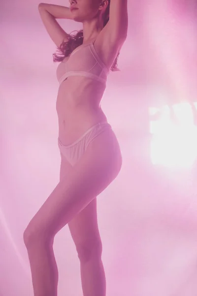 Cropped view of sexy girl in lingerie holding hands behind head while posing on pink and purple background with lighting — Stock Photo