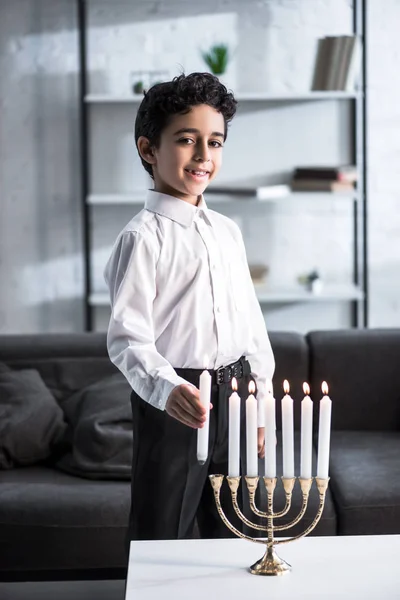 Lindo y sonriente chico judío en camisa sosteniendo vela - foto de stock
