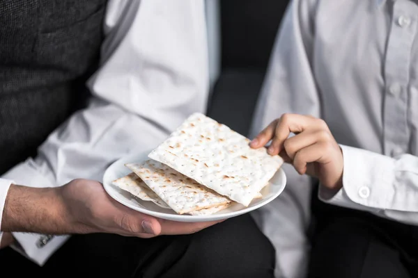 Vista cortada do pai judeu segurando placa e filho tomando matza no apartamento — Fotografia de Stock