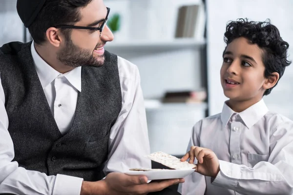 Sonriente padre judío sosteniendo plato e hijo tomando matza en apartamento - foto de stock