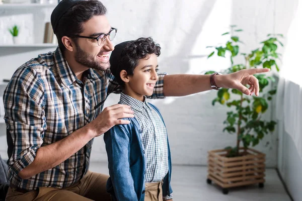 Smiling jewish father pointing with finger and looking through window with son in apartment — Stock Photo