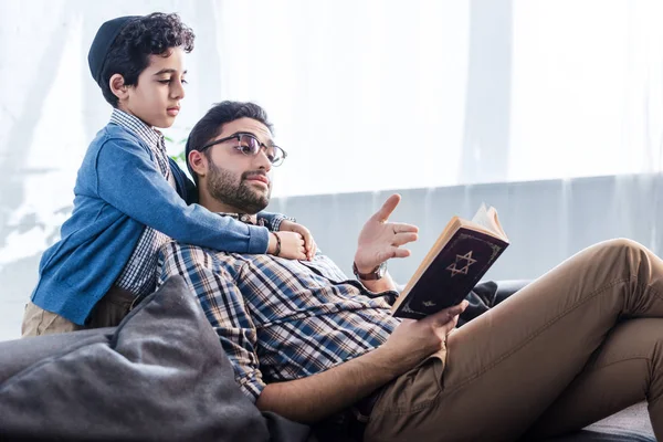 Jewish father pointing with hand at tanakh and talking with son in apartment — Stock Photo