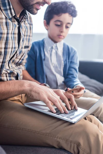 Visão cortada do pai judeu usando laptop e filho segurando cartão de crédito no apartamento — Fotografia de Stock
