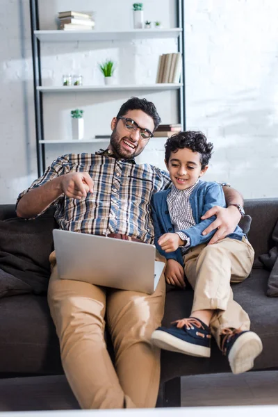 Sorrindo pai judeu apontando com o dedo para laptop e abraçando filho no apartamento — Fotografia de Stock