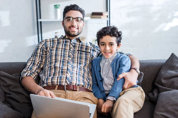 Sonriente padre judío con portátil e hijo mirando a la cámara en el apartamento - foto de stock